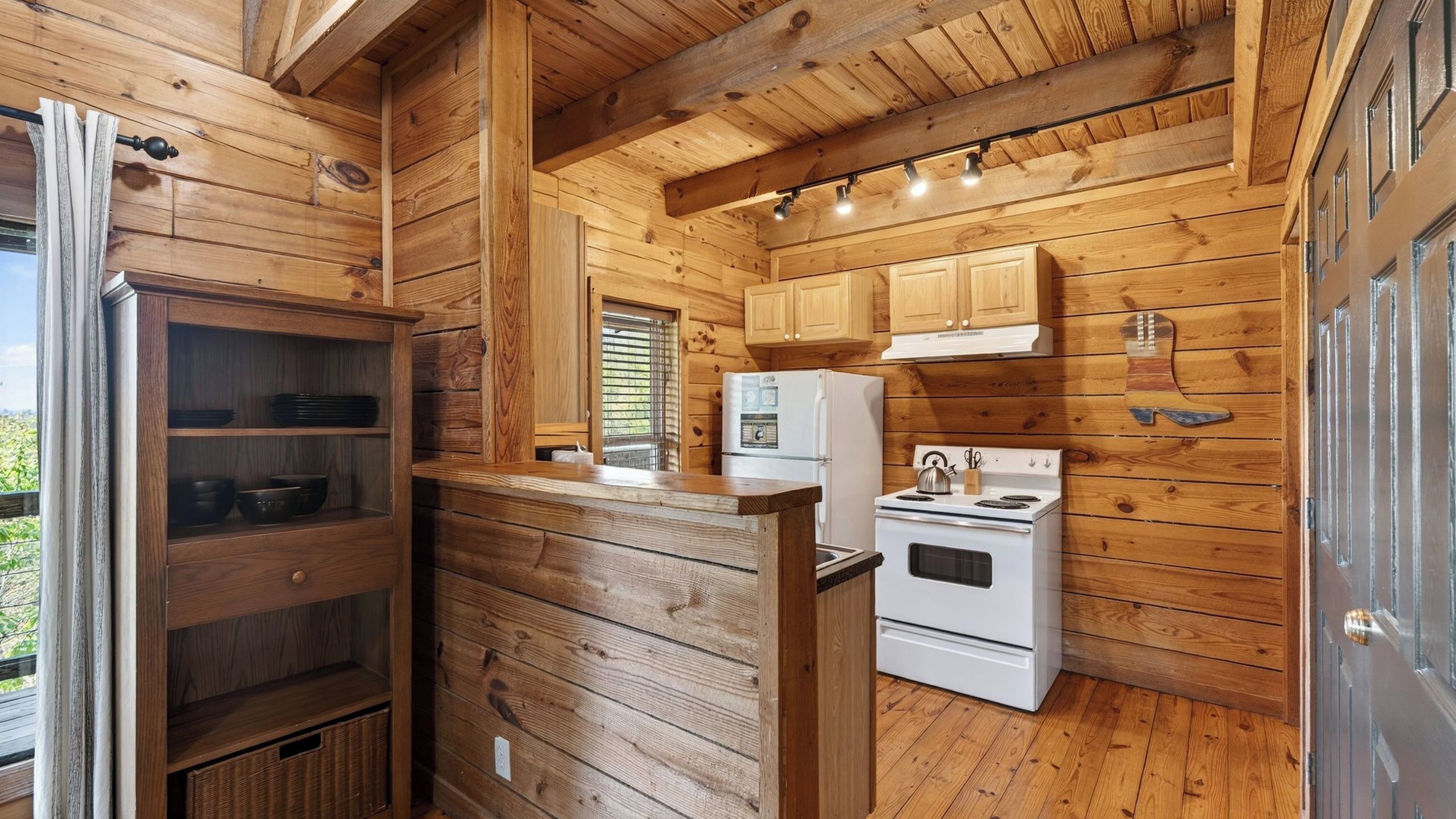 Interior de una cabaña de madera con zona de cocina, que incluye estufa, refrigerador y armarios de madera.