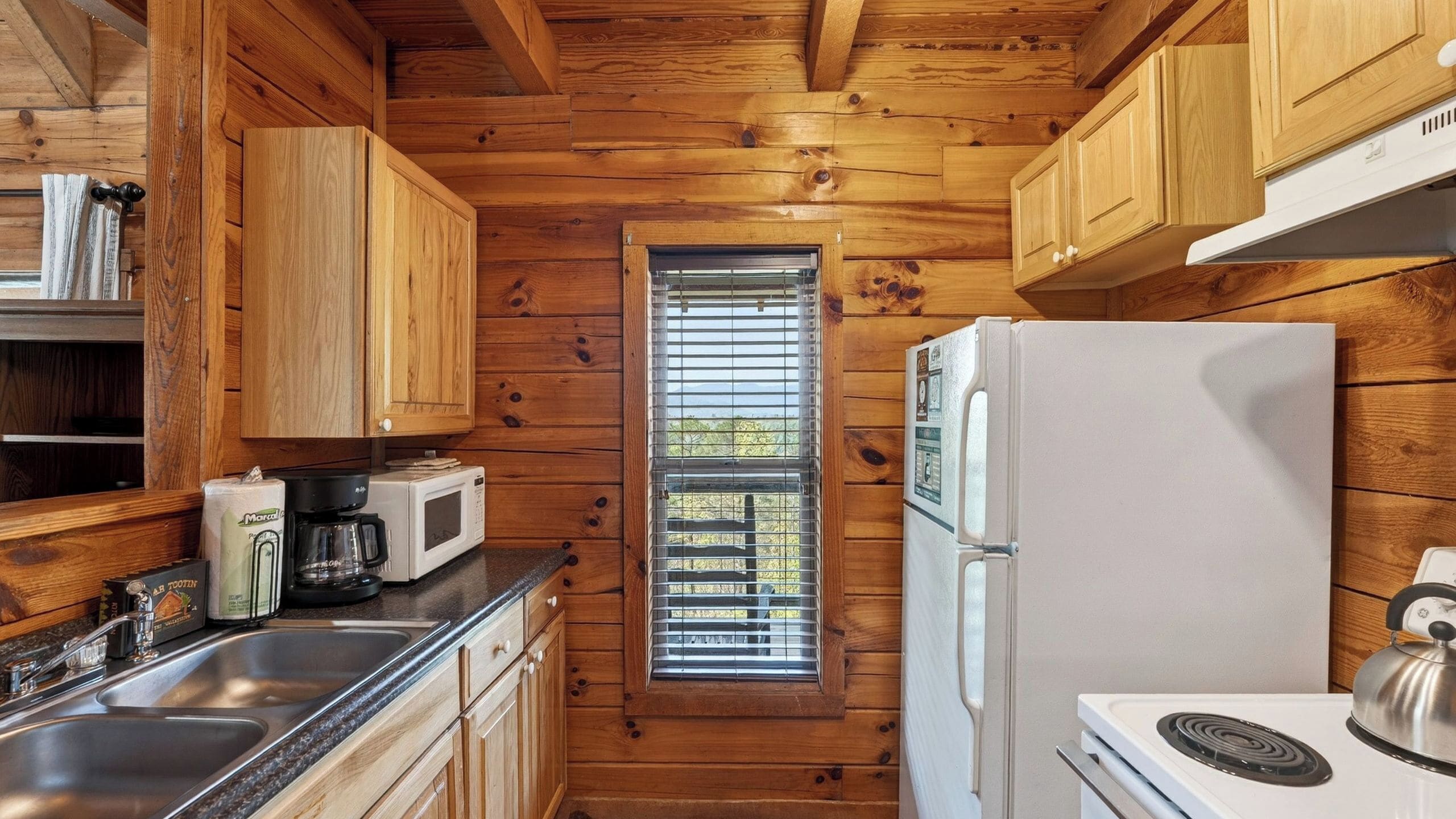Une cuisine compacte aux murs en bois, avec une fenêtre équipée de stores, un réfrigérateur blanc, une cuisinière et une machine à café.