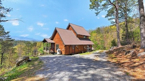 A log cabin with a gravel driveway, surrounded by trees and a clear sky.
