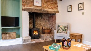 A living room with a brick fireplace, a television, a chair, a small table with board games, and a bottle of beer. - The Smithy's Cottage -  a cottage that sleeps 4 guests  in 2 bedrooms (Freston)