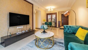 Ein modernes Wohnzimmer mit Fernseher, einem runden Couchtisch mit goldener Rahmung, einem grünen Sofa mit gelbem Kissen und einer Kronleuchter.