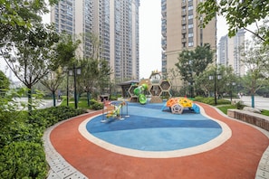 Un parque infantil con diversas estructuras de juego rodeado de follaje y altos edificios residenciales al fondo.