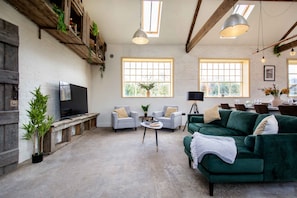 A spacious living room with a green sofa, two armchairs, a coffee table, and a TV. There are large windows, wooden beams, and a plant on the left.