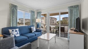 Ein Zimmer mit blauem Sofa, einem kleinen Tisch, einem Fernseher und einem Balkon mit Blick auf Gebäude.