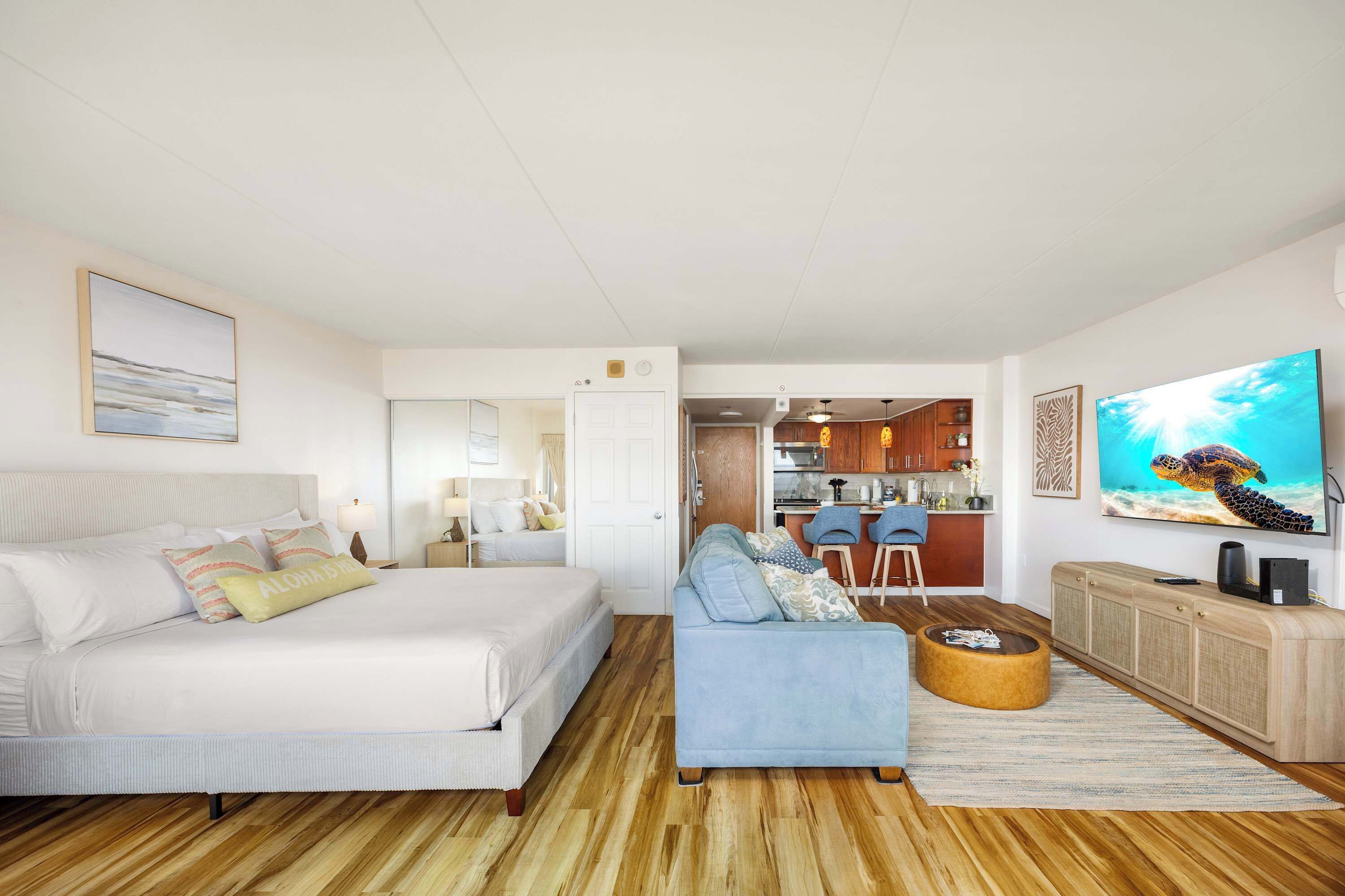 A bedroom with a bed, a sofa, a TV, and a kitchen in the background.