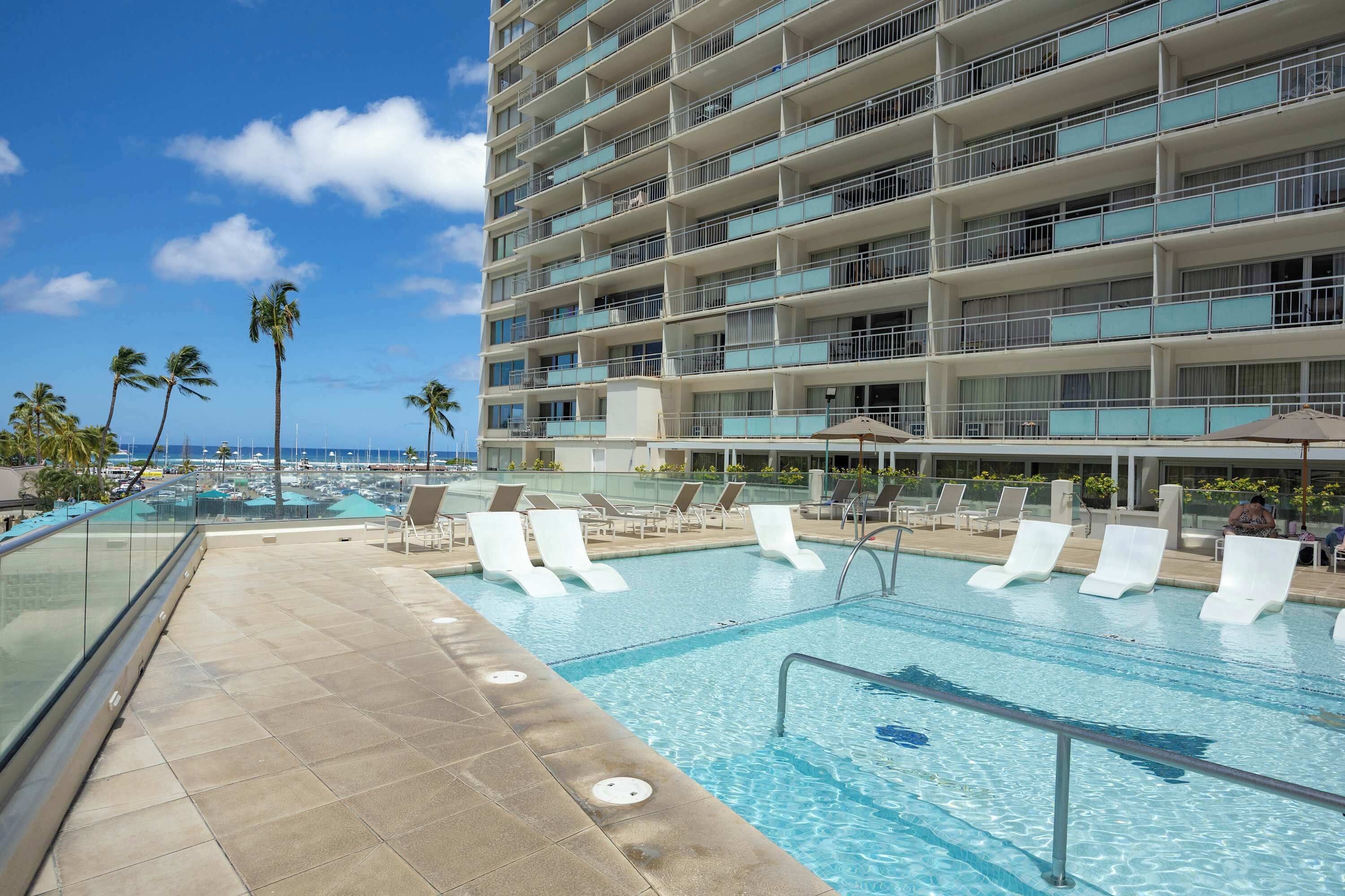 A hotel pool with clear blue water, white lounge chairs, and a view of the ocean.