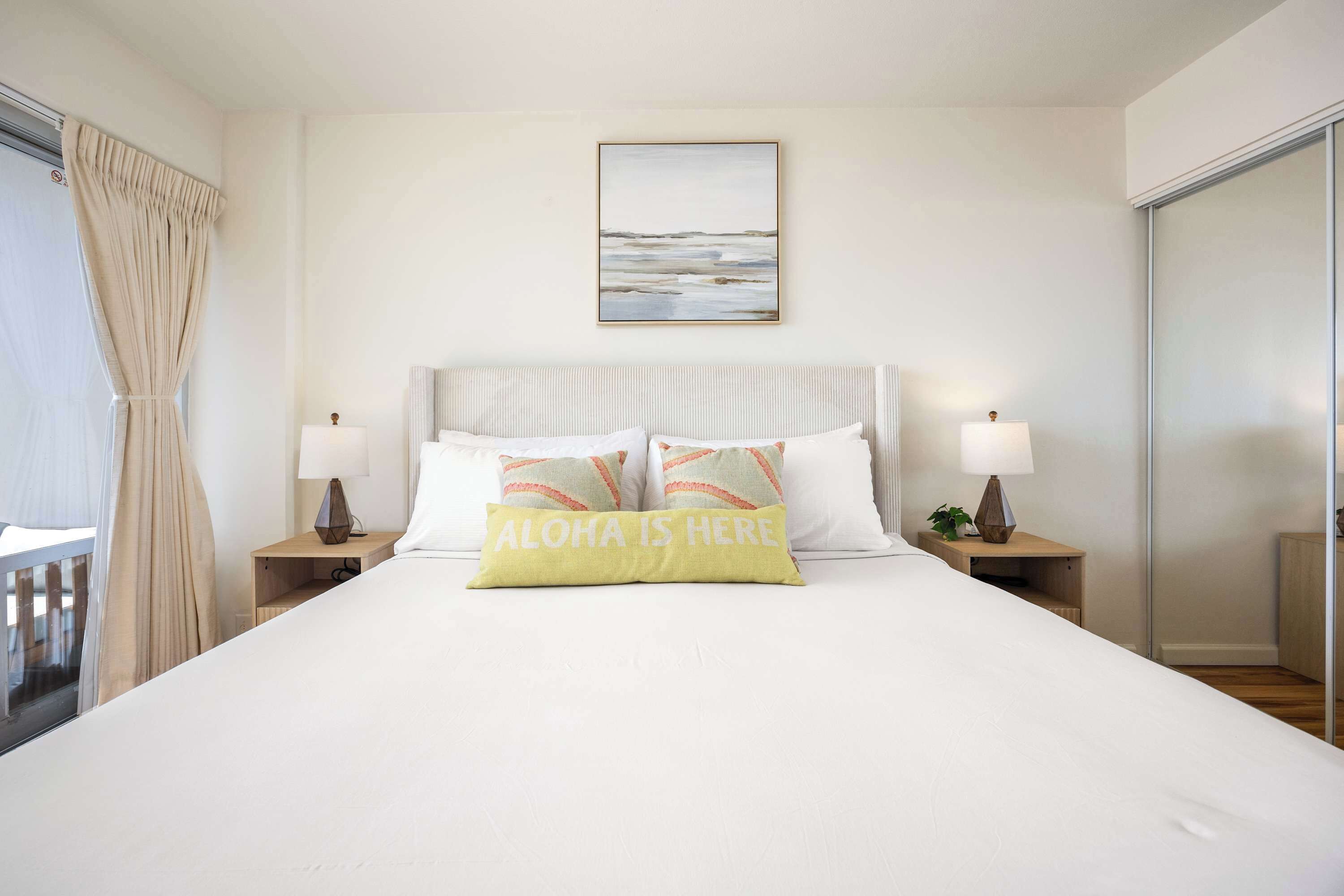 A hotel room with a large bed, two bedside tables with lamps, a view of the sea through the window, and a framed picture on the wall.