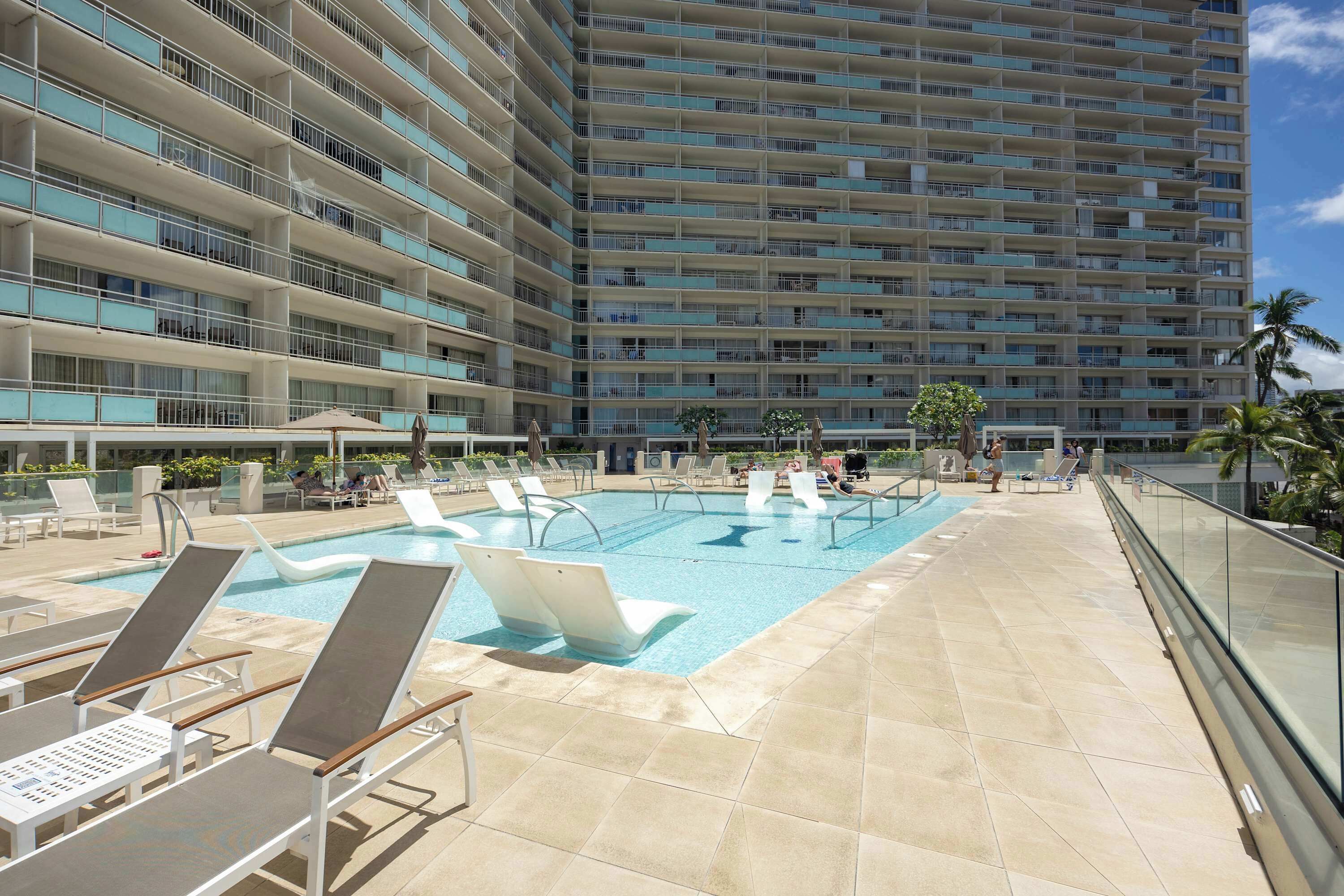 A hotel with a swimming pool, lounge chairs, and a tall building in the background.