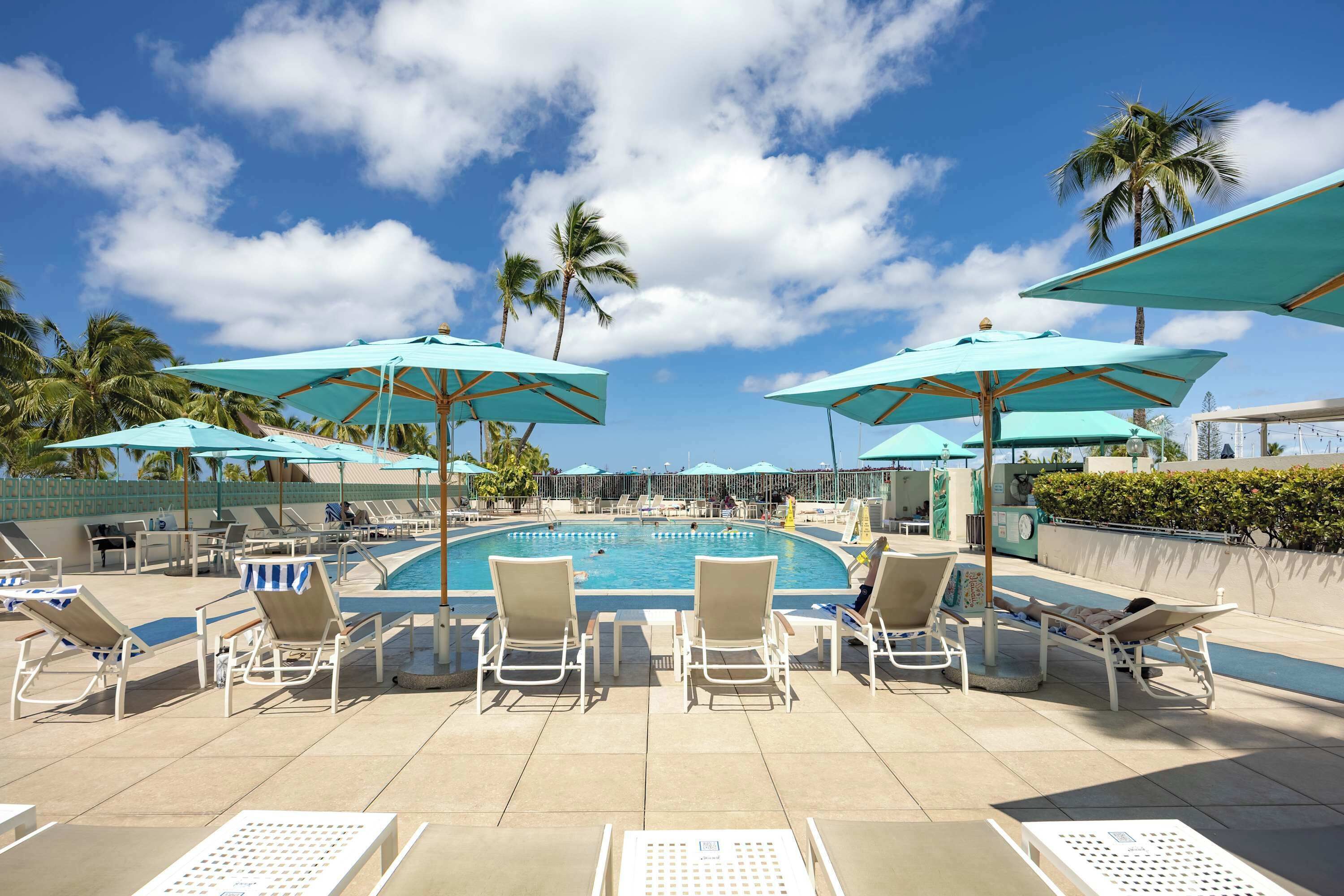 A poolside area with lounge chairs and umbrellas, a swimming pool, and palm trees.