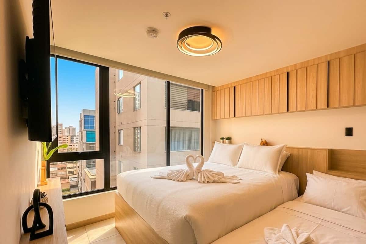 Una habitación de hotel moderna con una cama grande, un televisor montado en la pared y vistas de la ciudad a través de amplias ventanas.