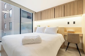 A modern hotel room with a large bed, a bedside table, a chair, and a view of the city through large windows. - Edificio Adagio by Host Ecuador (Quito)