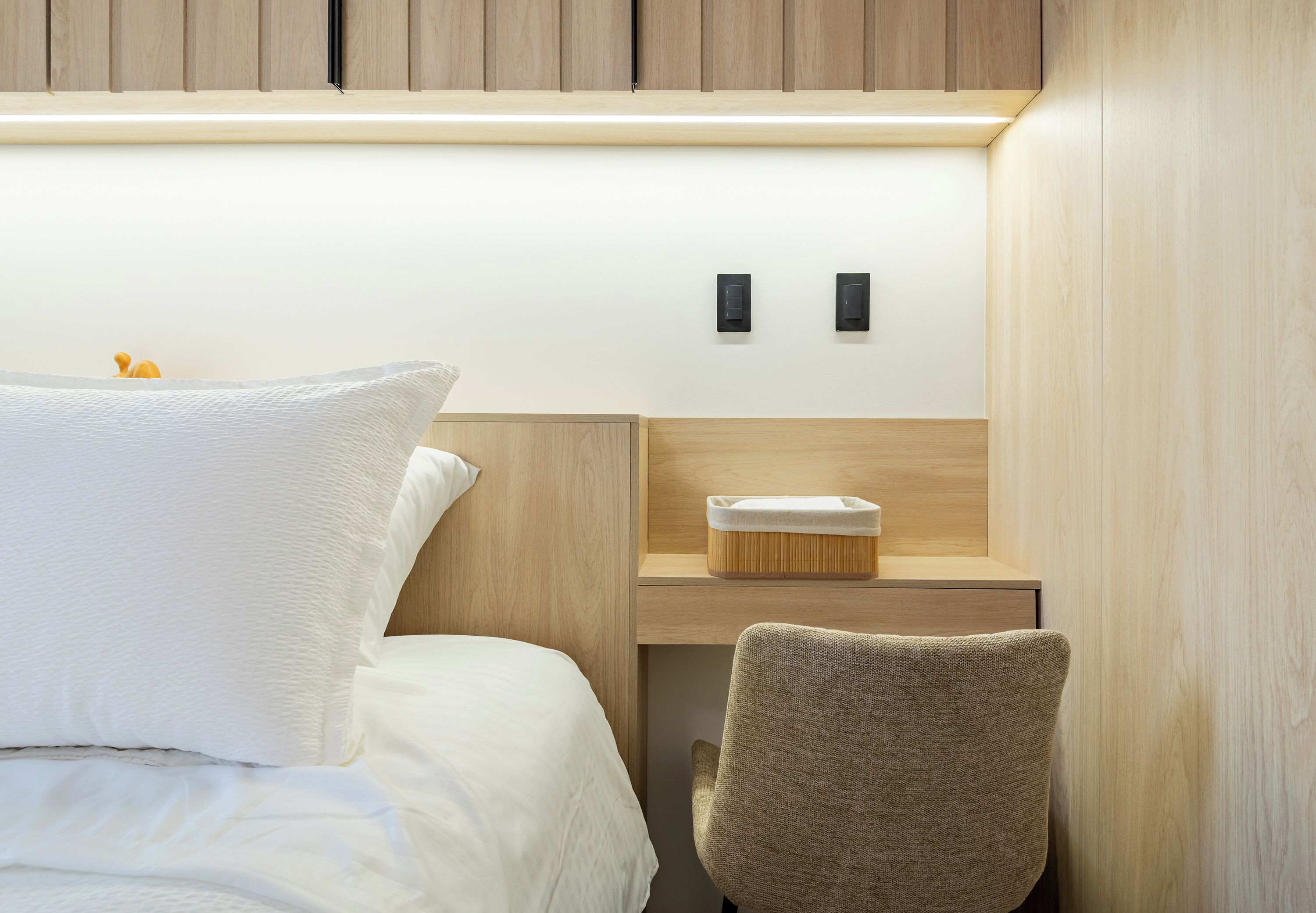 A hotel room with a bed, a chair, a desk with a lamp, and a small wooden shelf.