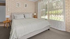 Un dormitorio con pared de ladrillo, una ventana grande con cortinas translúcidas, una cama con colcha, dos mesitas de noche con lámparas e impresiones botánicas enmarcadas en la pared.
