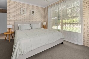 A bedroom with a brick wall, a large window with sheer curtains, a bed with a quilt, two bedside tables with lamps, and framed botanical prints on the wall. - Swimming Creek Beach House (Nambucca Heads)