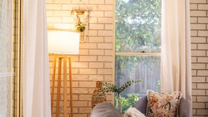 Una habitación acogedora con pared de ladrillo, una ventana con cortinas, una lámpara, una mesita con una planta y un sofá con un cojín estampado.