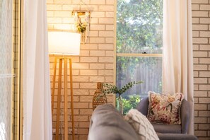 A cozy room with a brick wall, a window with curtains, a lamp, a small table with a plant, and a sofa with a floral pillow. - Swimming Creek Beach House (Nambucca Heads)