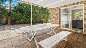 Un patio exterior con una mesa y bancos blancos, un muro de ladrillo y un área cubierta con vistas a follaje.