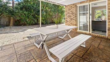 An outdoor patio with a white table and benches, a brick wall, and a covered area with a view of greenery.