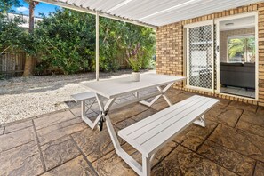 An outdoor patio with a white table and benches, a brick wall, and a covered area with a view of greenery. - Swimming Creek Beach House (Nambucca Heads)