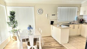 A modern kitchen with white cabinetry, a dining area with a white table and chairs, and a potted plant. - Cosy Villa - Pool Tennis & More | Pay 2 Stay 3 (Saint Georges Basin)