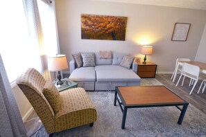 Ein Wohnzimmer mit einer Couch, einem gemusterten Sessel, einem Couchtisch, einem Gemälde an der Wand und einem kleinen Tisch mit einer Lampe.