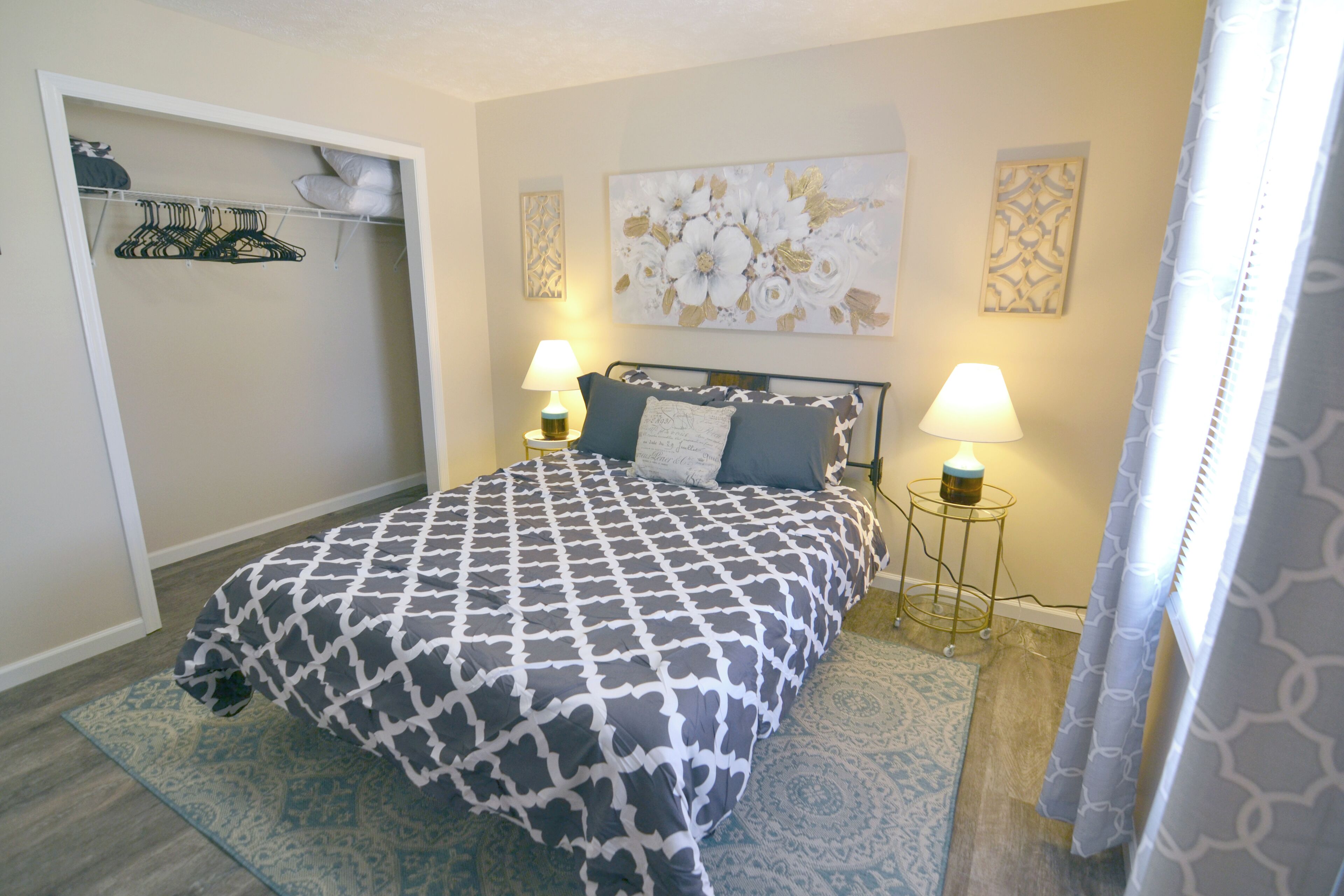 A bedroom with a bed, two bedside lamps, a wall art piece, and a closet with hanging items.
