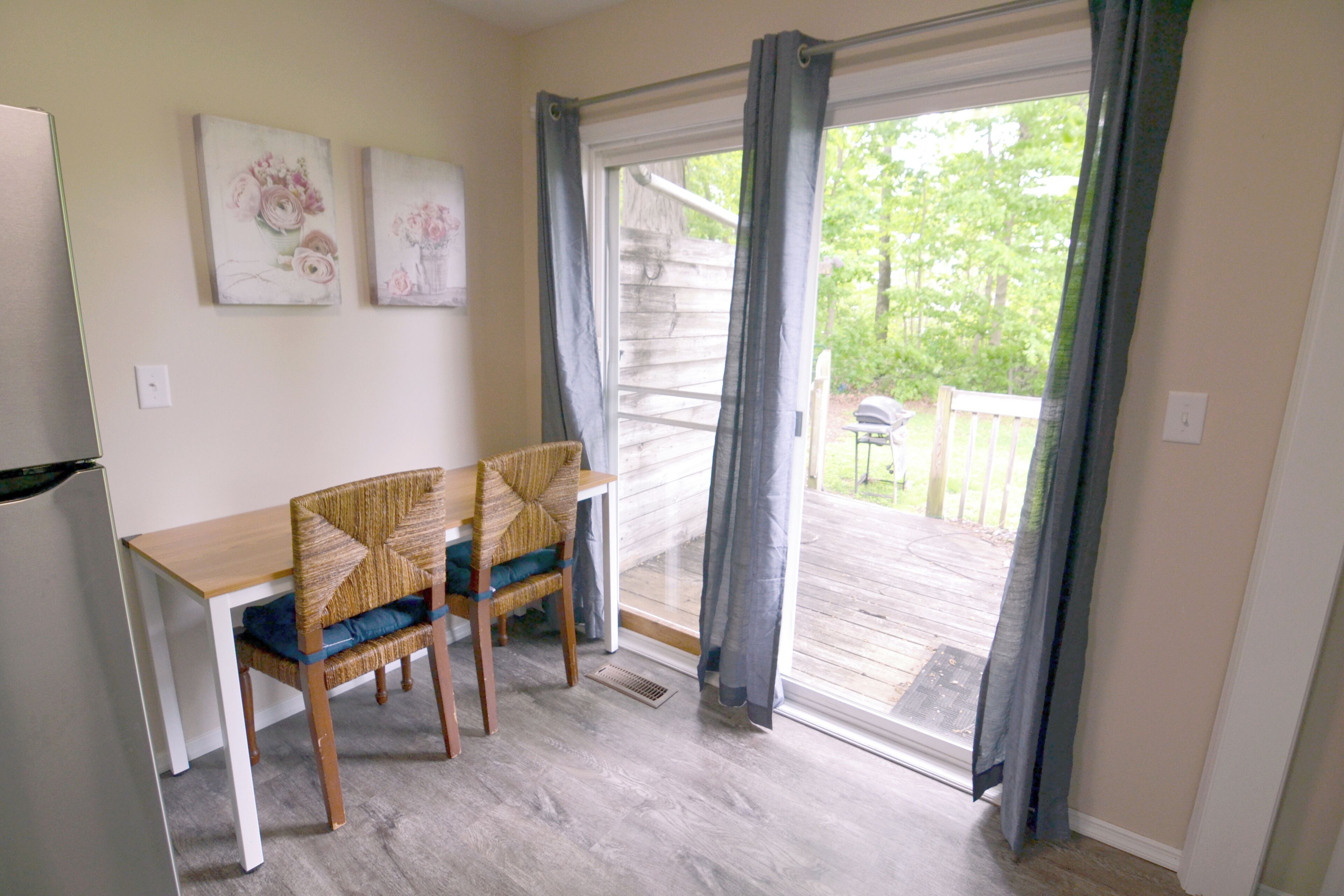 A room with a wooden table and two chairs, a window with curtains, and two wall-mounted floral paintings.