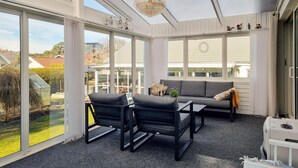 Une véranda avec de grandes fenêtres, un canapé, deux fauteuils, un lustre et une vue sur l’extérieur.