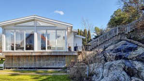 Une maison moderne dotée de grandes fenêtres, d’une terrasse en bois et d’un mur de soutènement en pierre.