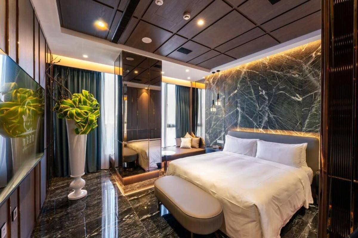 A modern hotel room with a large bed, a seating area, a mirror, and a large plant.