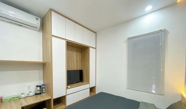 Une chambre avec un lit, un téléviseur posé sur un meuble en bois, une fenêtre avec des stores et une étagère contenant des articles de toilette.