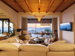 A spacious living area with a large sofa, a dining table, and chairs. There is a television mounted on the wall and a view of the sea through large windows. - Illume Mykonos | Villa with infinity pool sea view (Mykonos)