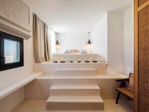 A bedroom with a bed, two bedside lamps, a window with curtains, and a rattan chair. - Illume Mykonos | Villa with infinity pool sea view (Mykonos)