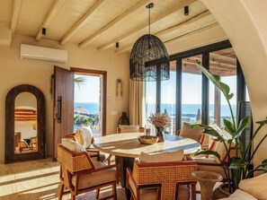 A room with a round table, wicker chairs, a large window with a view of the sea, and a hanging light fixture. - Illume Mykonos | Villa with infinity pool sea view (Mykonos)