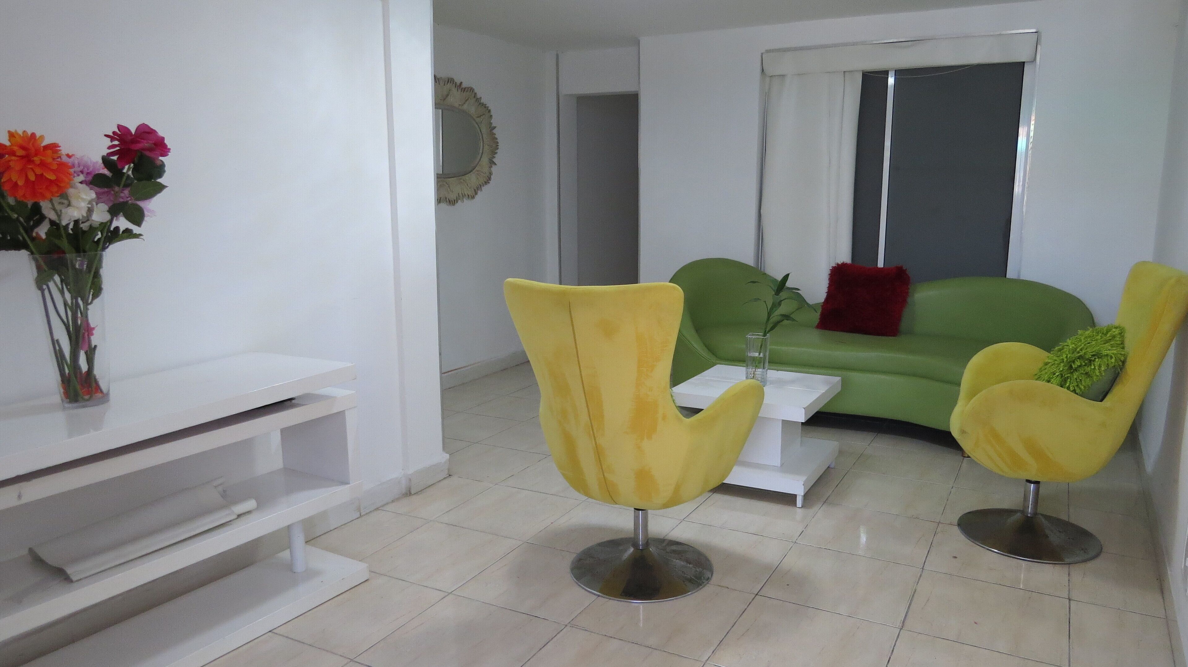 A modern living room with a green sofa, yellow chairs, a white coffee table, and a vase of flowers on a white console table.