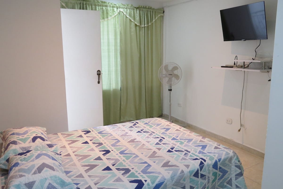 Un dormitorio con una cama, un ventilador de pie, un televisor montado en la pared y una puerta con cortina.