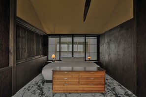Quarto com cama, cómoda de madeira, duas mesas de cabeceira com abajures e janela com persianas verticais.