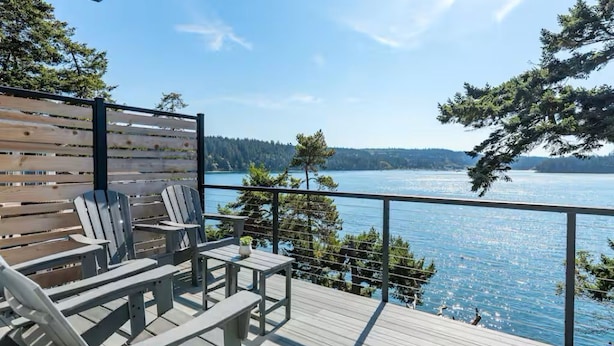 A deck with Adirondack chairs and a table overlooking a lake with forested hills in the background.