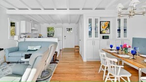 A spacious living area with a sofa, dining table, and chairs, a kitchen with white cabinets, and a hallway leading to another room.