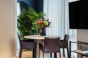 Un espace repas moderne avec une table ronde, quatre chaises, un vase avec des fleurs, une grande plante en pot et un téléviseur à écran plat fixé au mur.