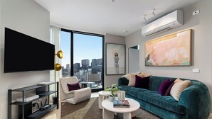 A modern living room with a city view, a flat-screen TV, a teal sofa, and a coffee table. - Skyline Suite Near Crown · Panoramic Yarra Views. Heated Pool, Gym, Sauna, Steam (Southbank)