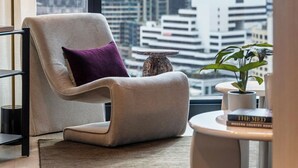 A modern living room with a city view, a comfortable chair, a small table, and a potted plant. - Skyline Suite Near Crown · Panoramic Yarra Views. Heated Pool, Gym, Sauna, Steam (Southbank)