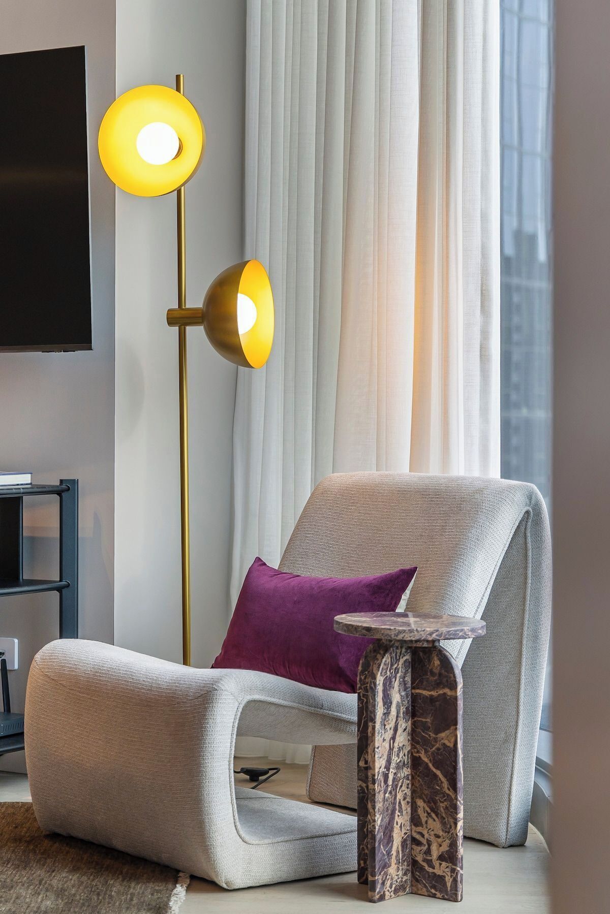 Una sala de estar moderna con un sofá gris, una almohada morada, una mesita de noche de mármol y una lámpara de pie con base dorada.