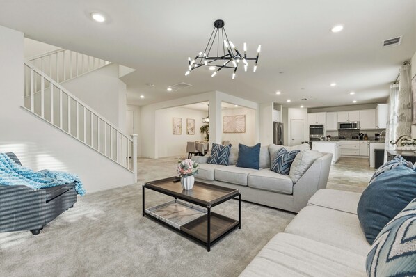 A spacious living room with a staircase, a modern chandelier, a sofa set, and a kitchen area.