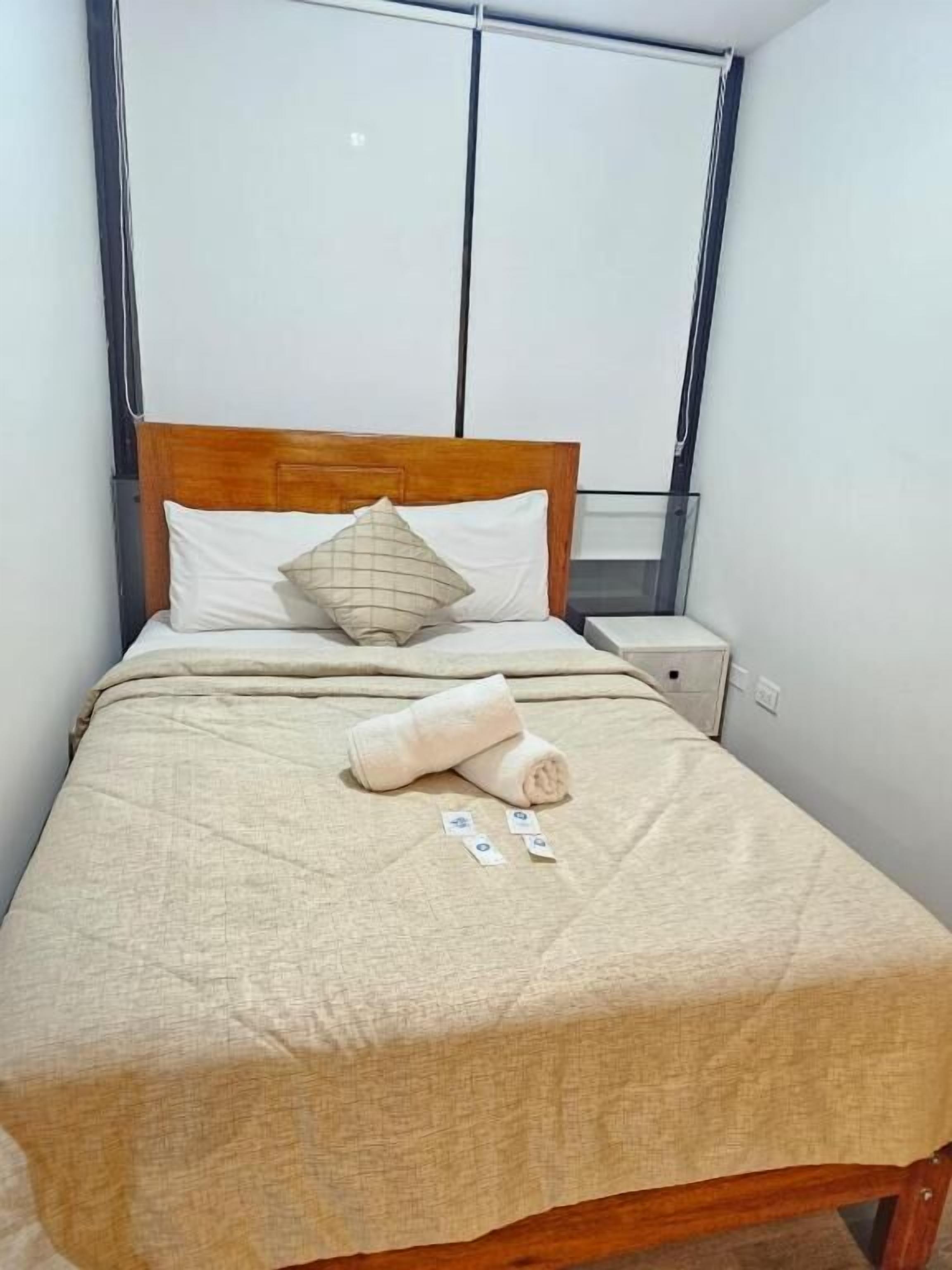 Un lit bien fait, avec une tête de lit en bois, une table de chevet et une fenêtre équipée de stores.