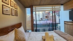 Habitación de hotel con techo de madera, una cama grande con cabecera, mesita de noche, televisor y balcón con vistas a edificios.
