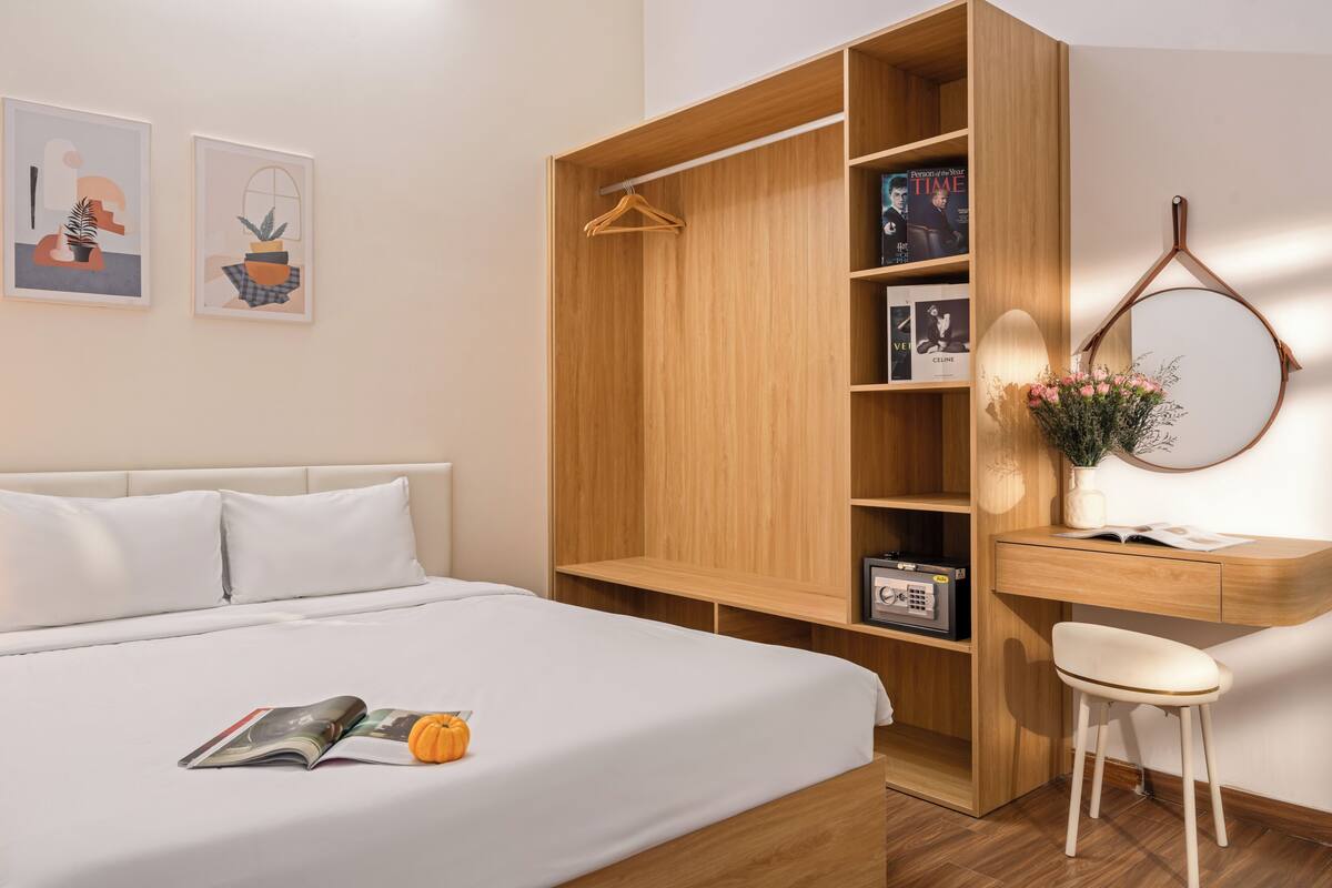 A bedroom with a bed, a wooden wardrobe, a desk with a chair, and a mirror.