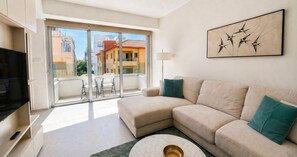 A modern living room with a beige sofa, a flat-screen TV, a glass door leading to a balcony, and a cityscape view. - Victoria Levanco Apartment (Limassol)