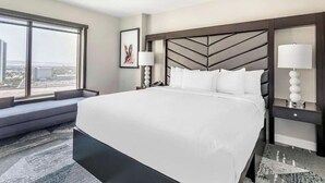 Una camera d'albergo con un letto grande, vista sulla città attraverso una finestra, un divano e un comodino con una lampada.