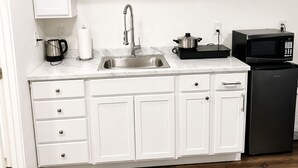 A kitchen with white cabinets, a stainless steel sink, a kettle, a microwave, and a black refrigerator.